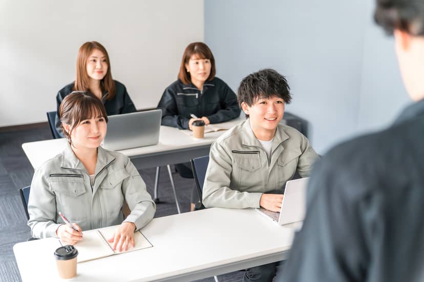 作業着の社員が研修室で説明を聞いている場面