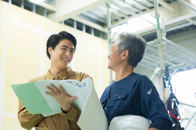 内装工事中の現場で、図面を手にした若手職人とヘルメットを抱えたベテランが笑顔で話している