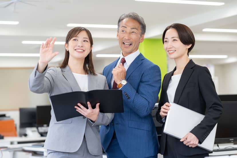 事務所でバインダーやノートパソコンを手に、笑顔で並ぶスーツ姿の社員3名