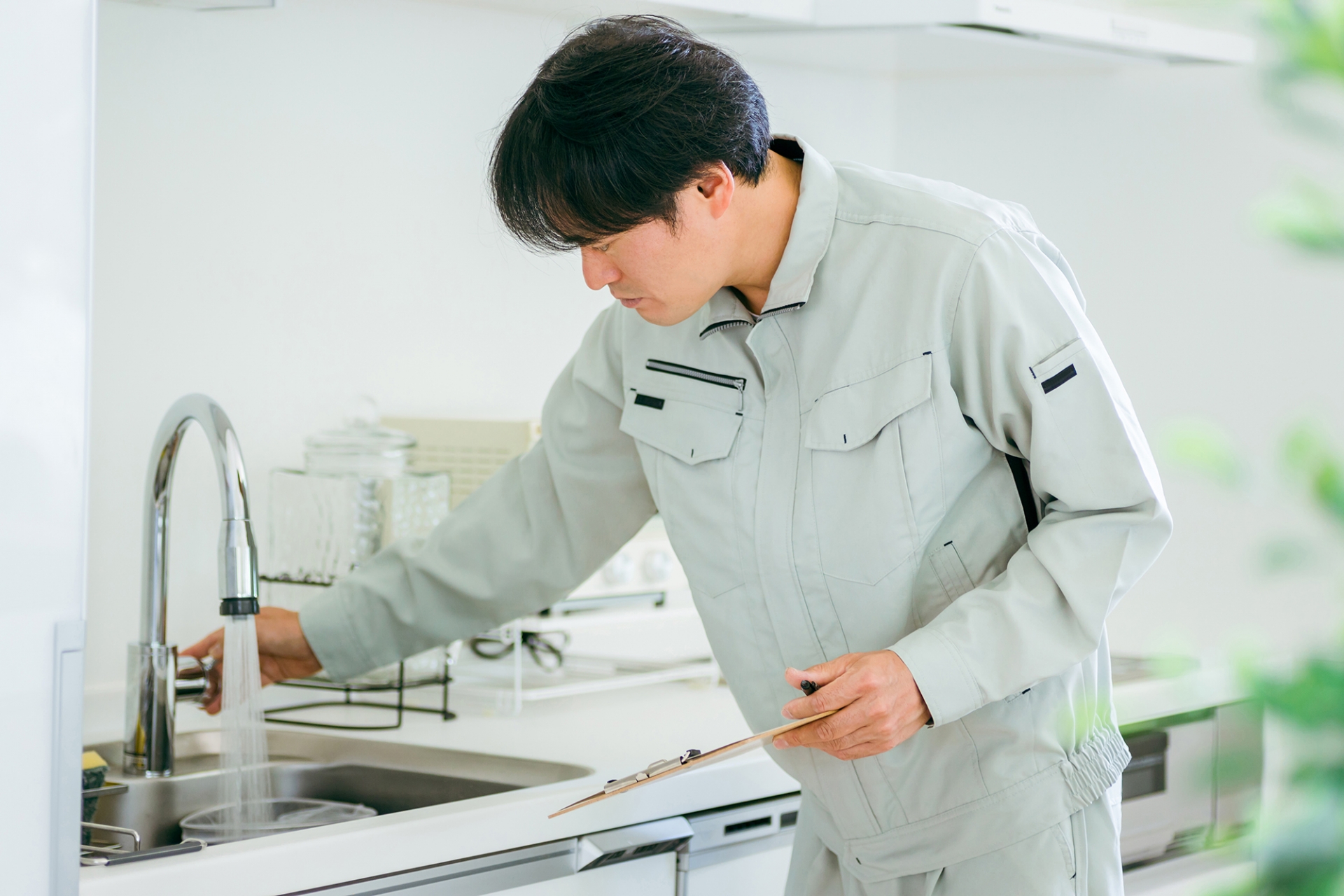 グレーの作業着の男性がキッチンのシンクの前でかがみ、水栓に触れながらバインダーの用紙に記入している写真