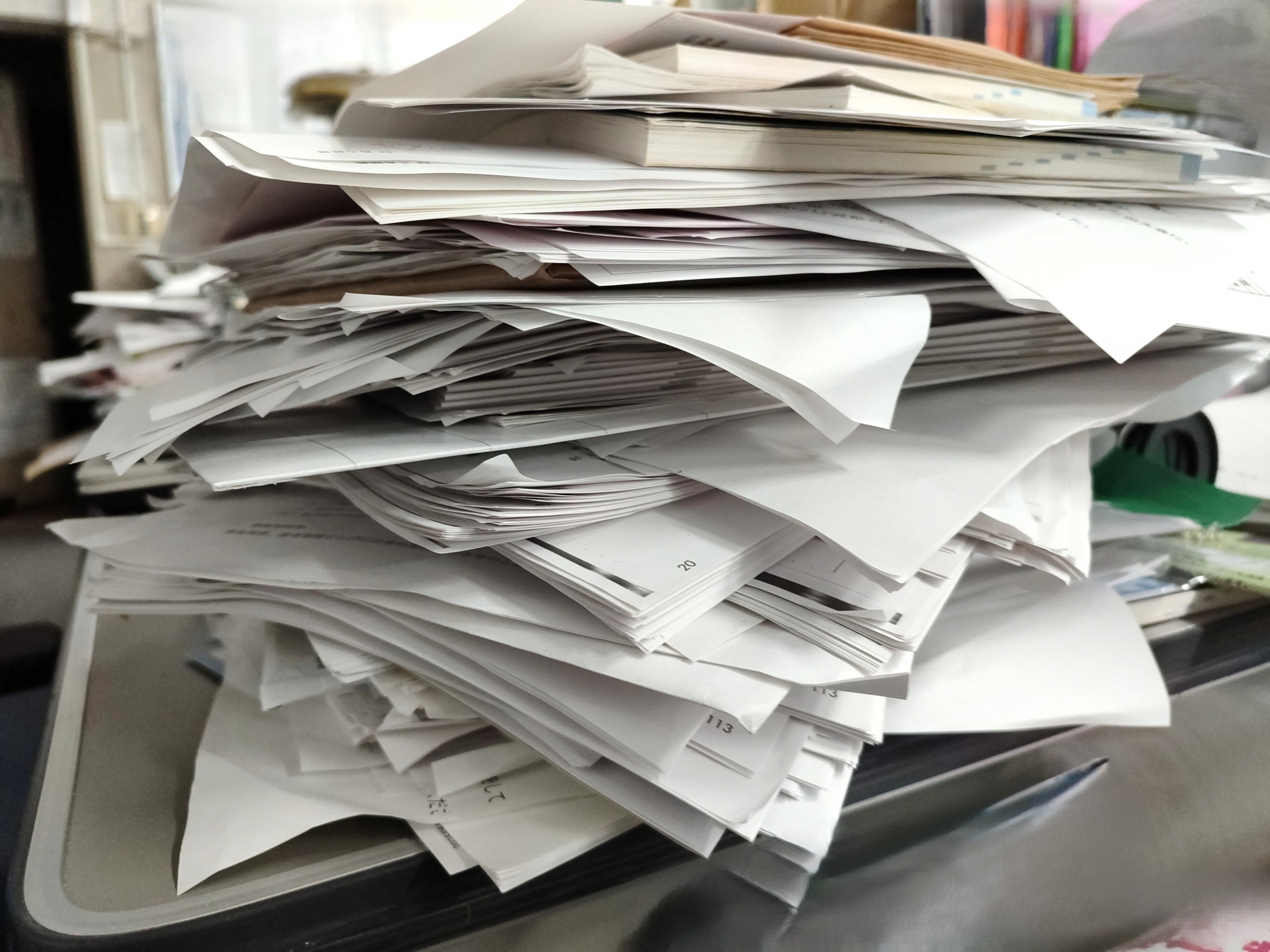 事務所の机の上に、プリントした書類が付箋やクリップを挟んだまま乱雑に高く積み上がっている写真