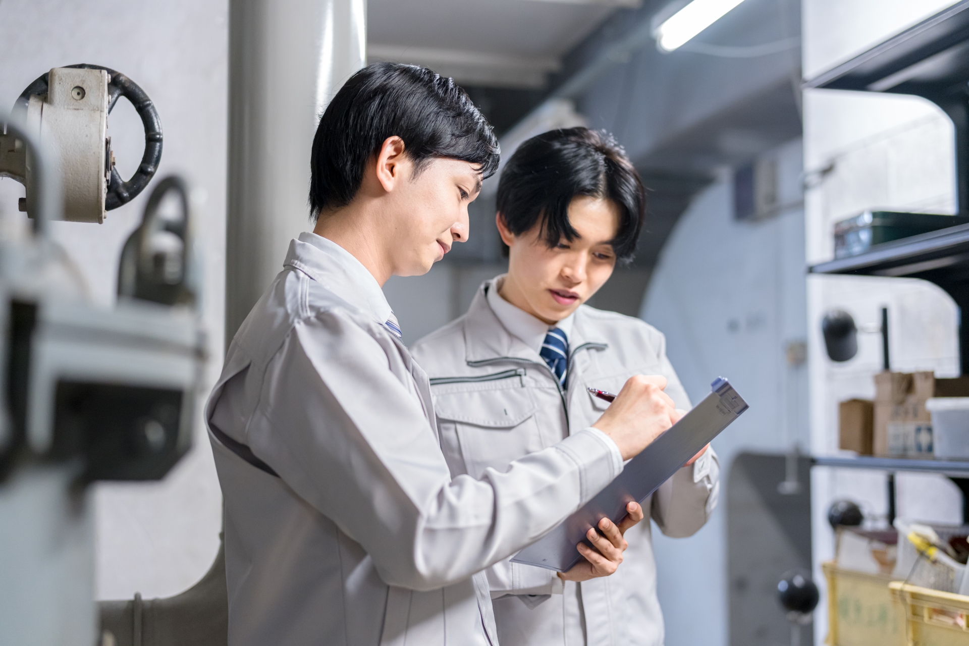 倉庫のような場所で、作業着の二人がクリップボードに書き込んでいる横顔の写真。背景の棚に部材が並んでいる