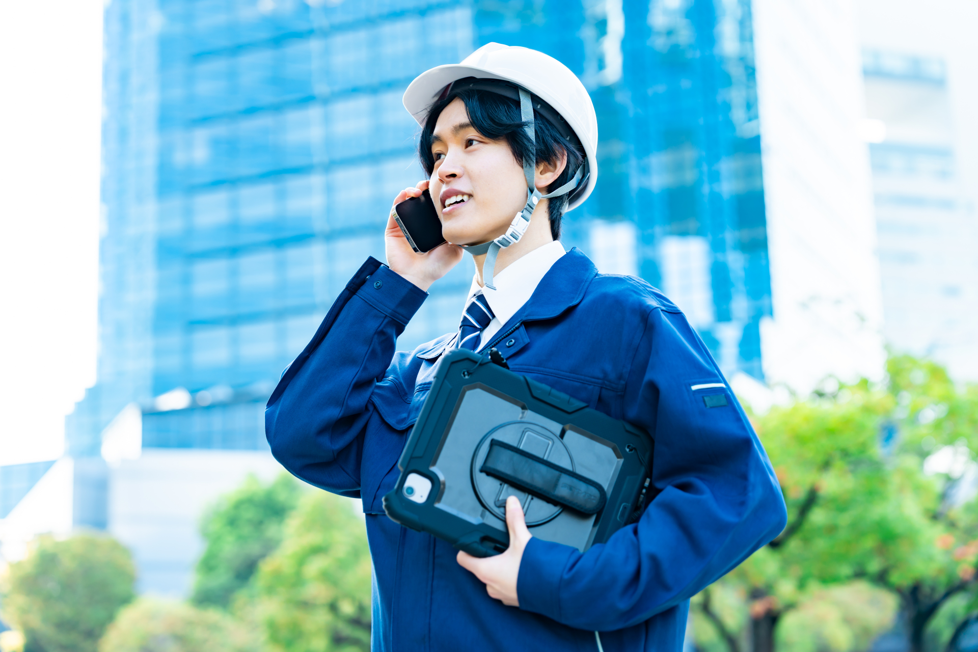濃紺の作業着に白いヘルメットをかぶった男性が、屋外でスマートフォンを耳に当てて話しながら、黒いタブレットを小脇に抱えている写真