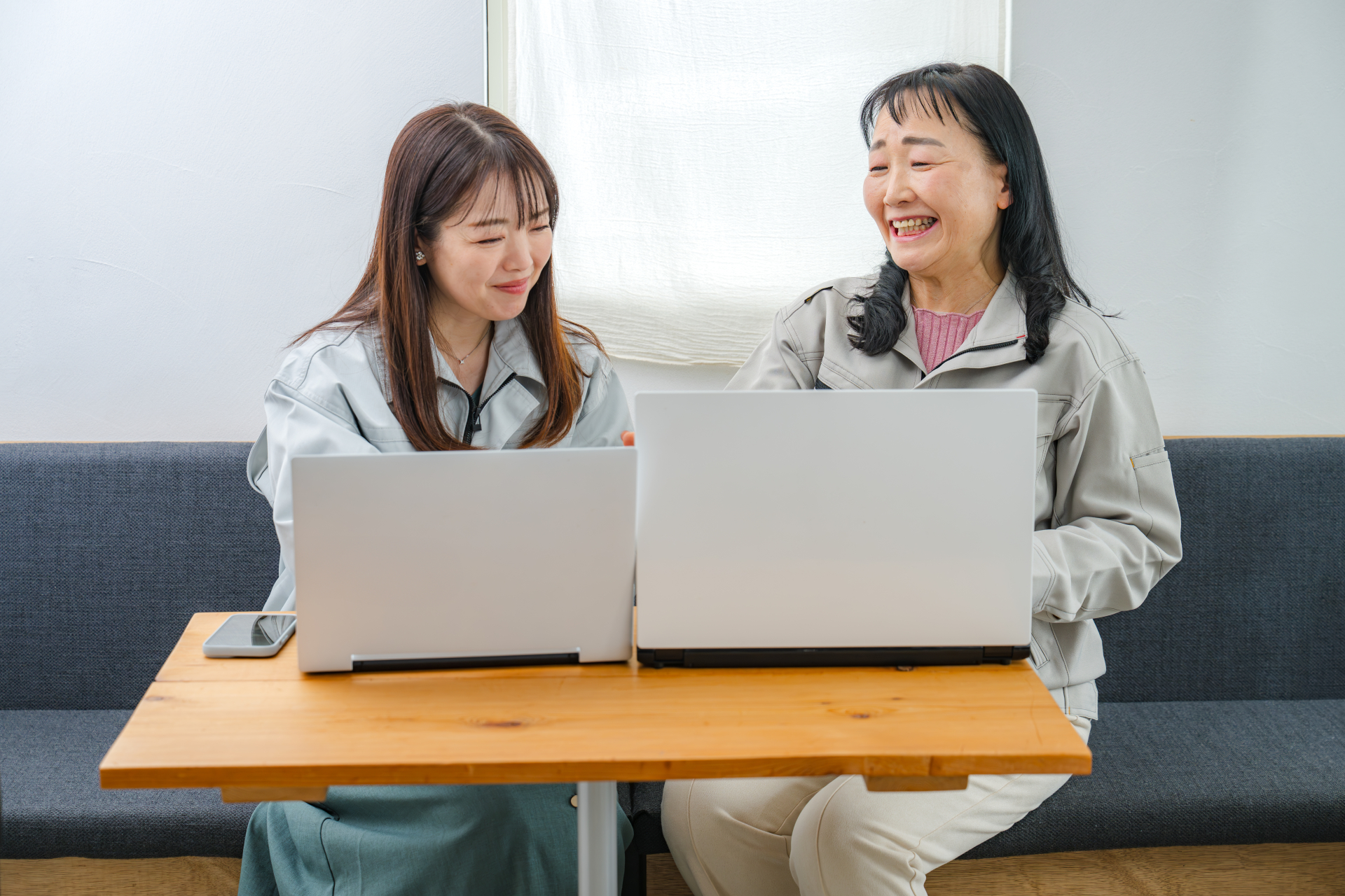 作業着姿の年配と若手の女性2名が、ソファに座って木のローテーブルにノートパソコンを2台並べ、画面を一緒に見ている写真