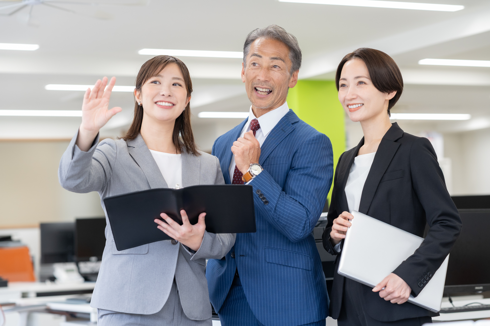 事務所でバインダーやノートパソコンを手に、笑顔で並ぶスーツ姿の社員3名