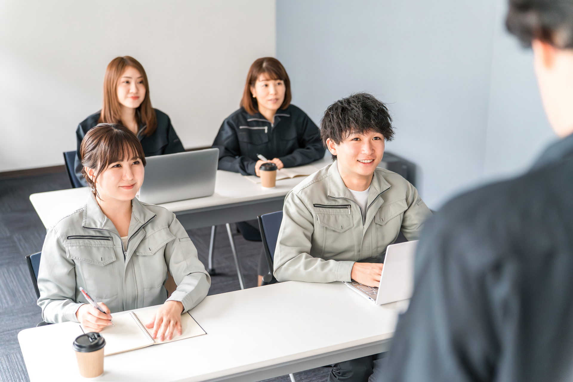 作業着の社員が研修室で説明を聞いている場面