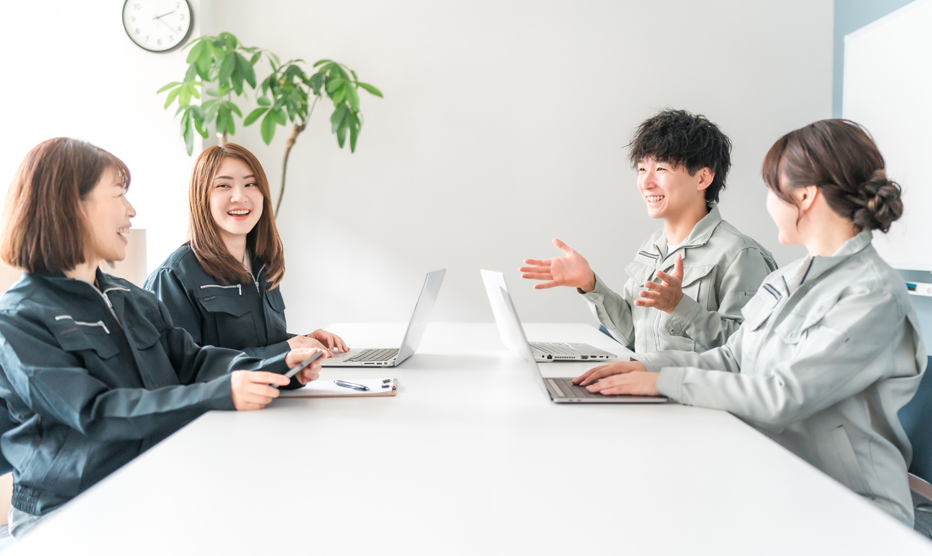 白い会議テーブルを挟んで、濃紺の作業着の二人とライトグレーの作業着の二人が向かい合い、ノートパソコンを前に話している写真