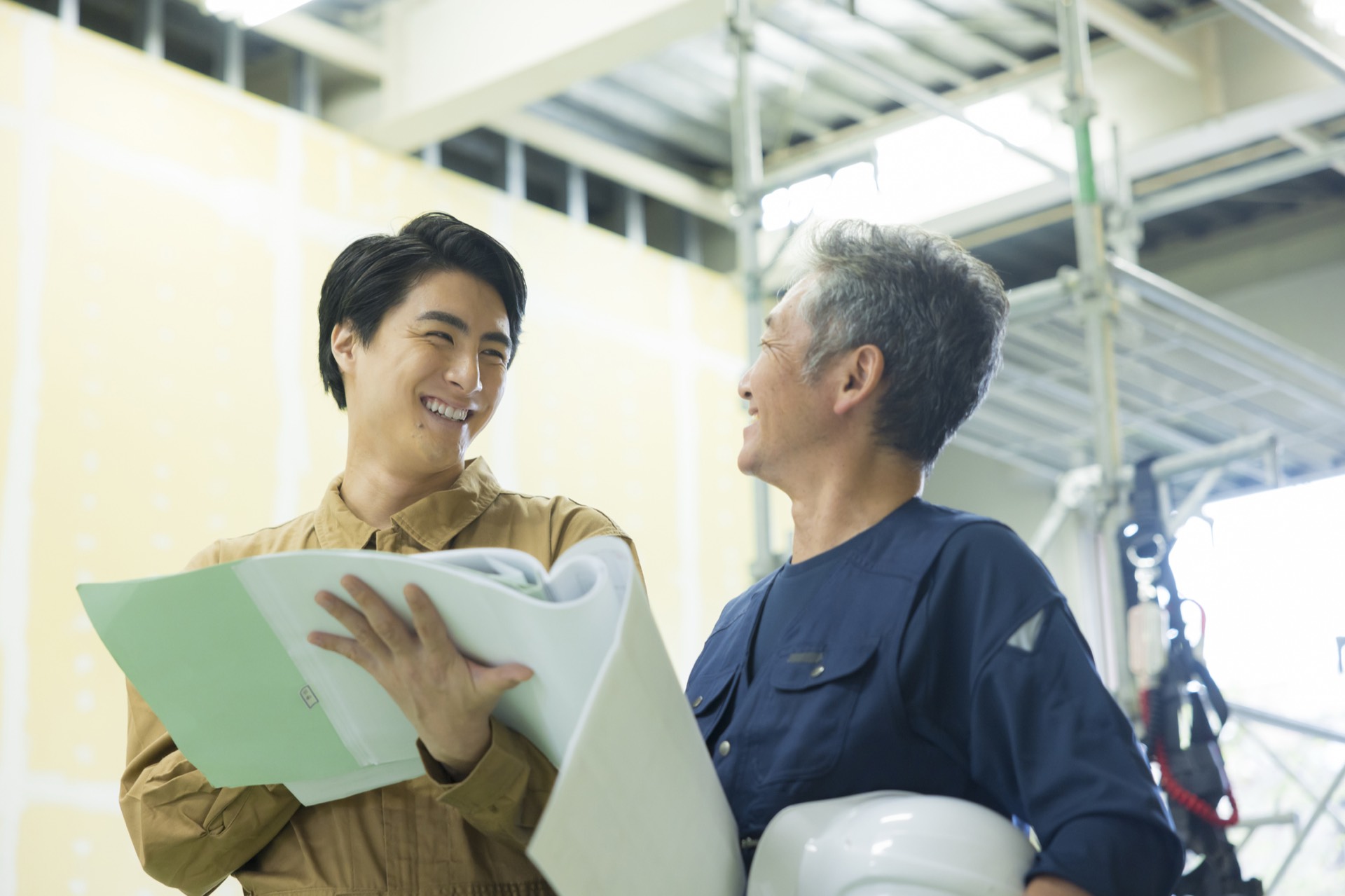 内装工事中の現場で、図面を手にした若手職人とヘルメットを抱えたベテランが笑顔で話している